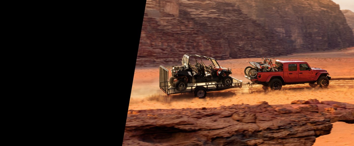 The 2021 Jeep Gladiator Rubicon being driven off-road with two dirt bikes in the truck bed and towing a trailer with a UTV in it.